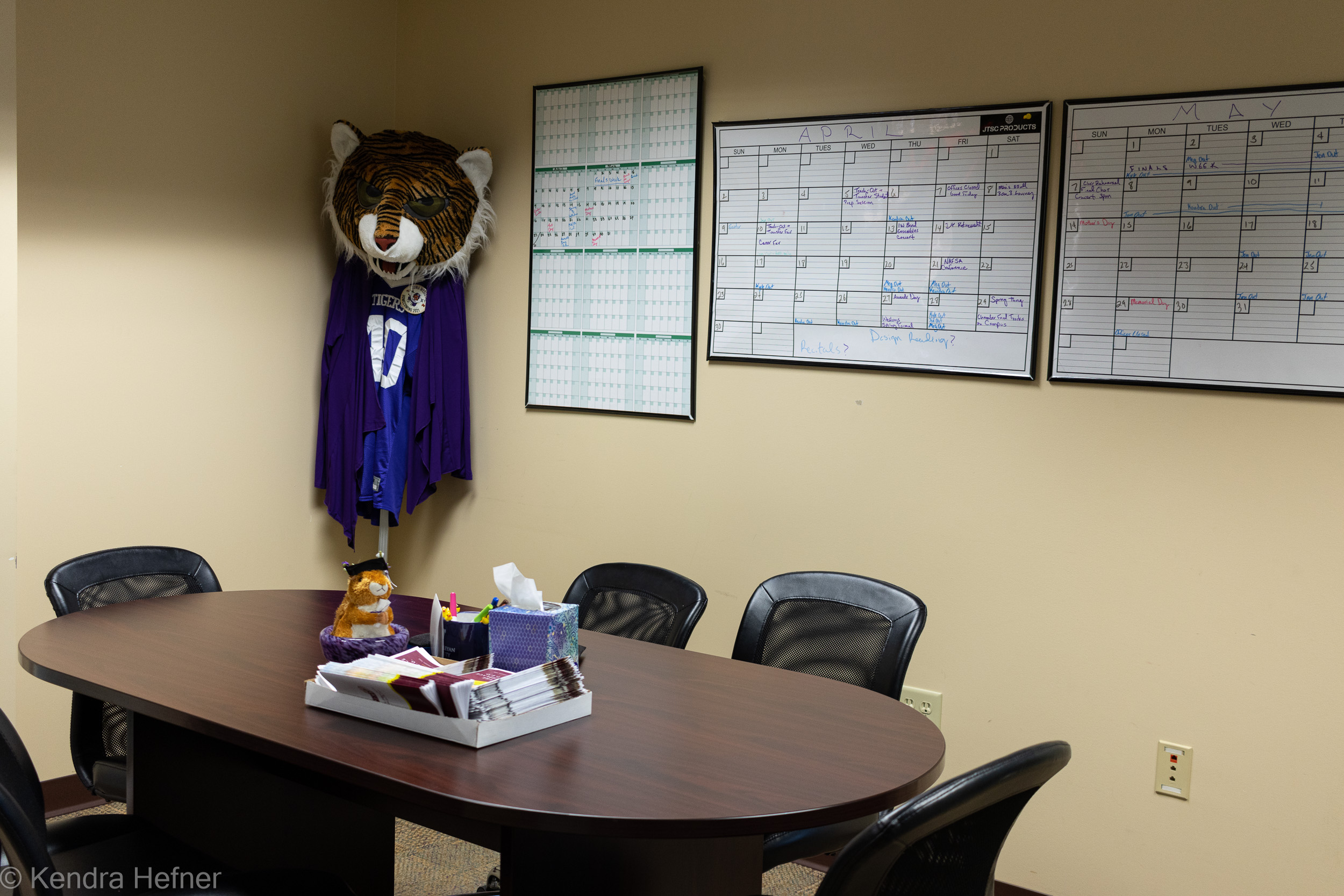 A conference table surrounded by empty chairs, Kevin (the marketing mascot) in the corner, monthly calendars for April and May hang on the wall. The annual calendar is empty except for April and May. Spring 2023.