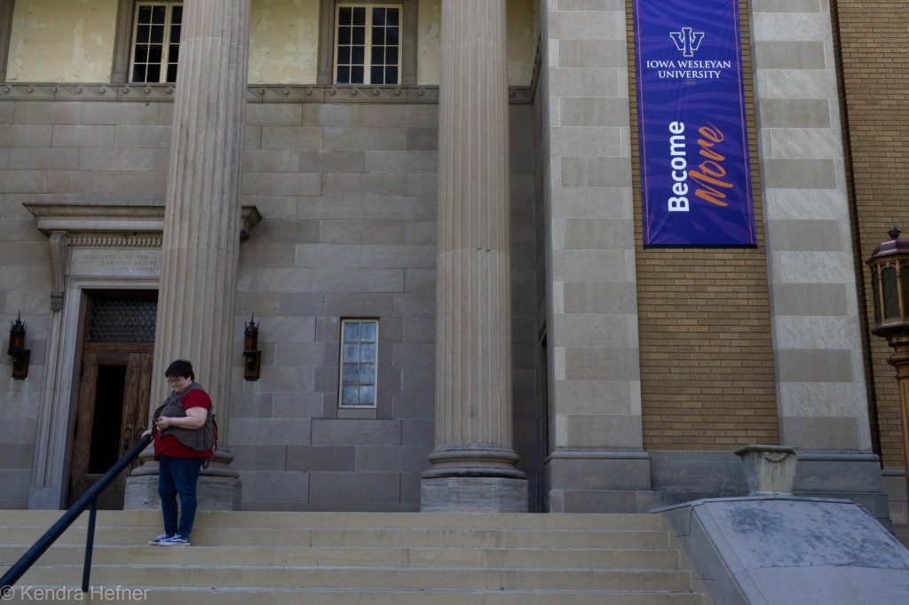 Jen Koch, IW graphic designer, on the stairs to the P.E.O. Memorial Library. "Become More" Admissions banner hangs on building outer wall. Spring 2023.