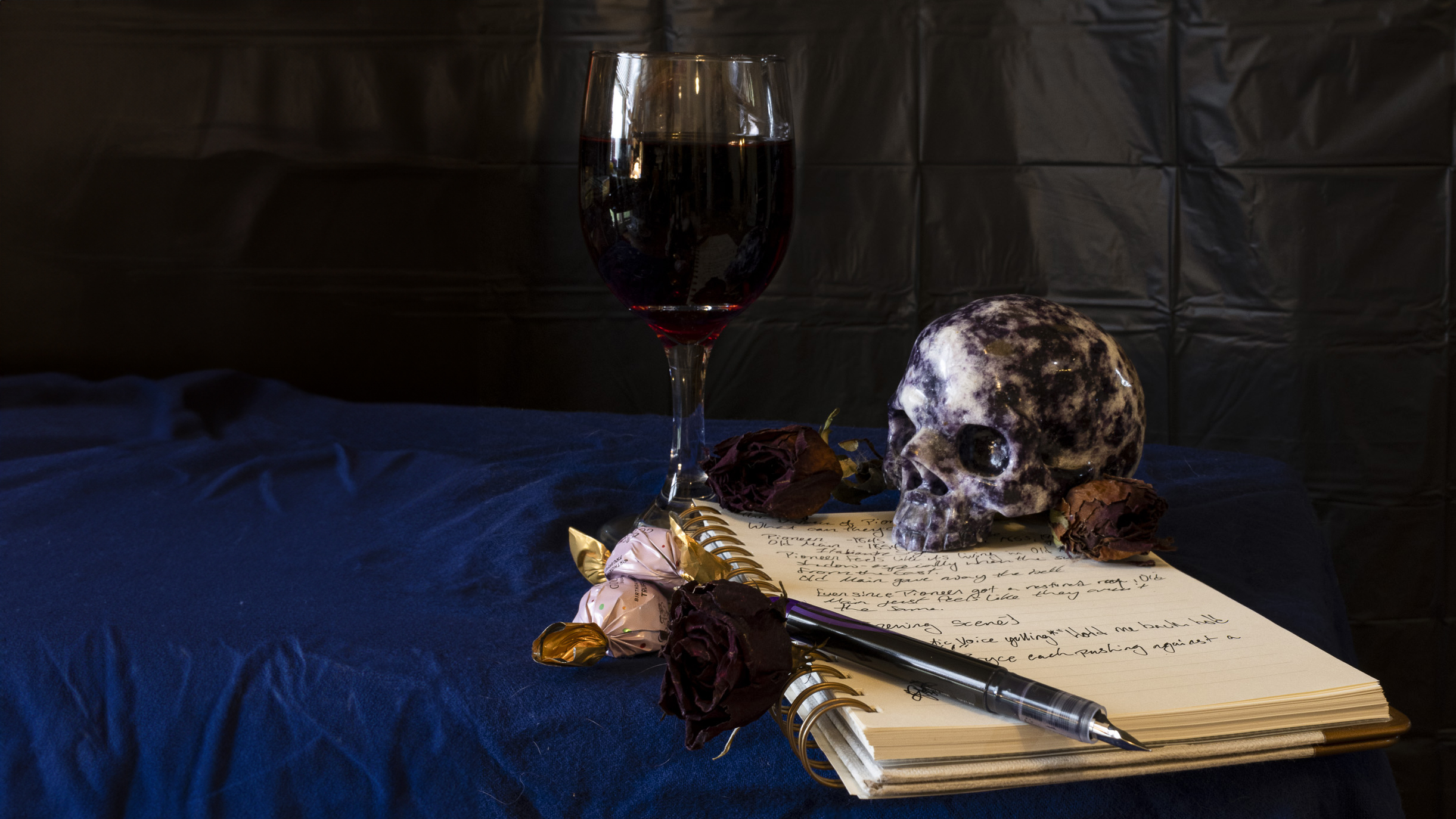 Still life image featuring filled wine glass, a purple rock in the shape of a skull, a note pad, a pen, and flowers.